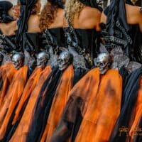 Desfile de personas disfrazadas con calaveras y capas naranjas.