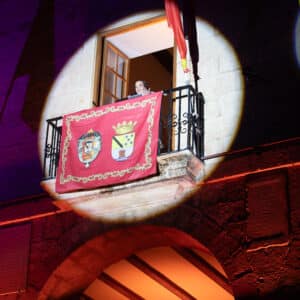 Persona en balcón iluminado con estandarte rojo.